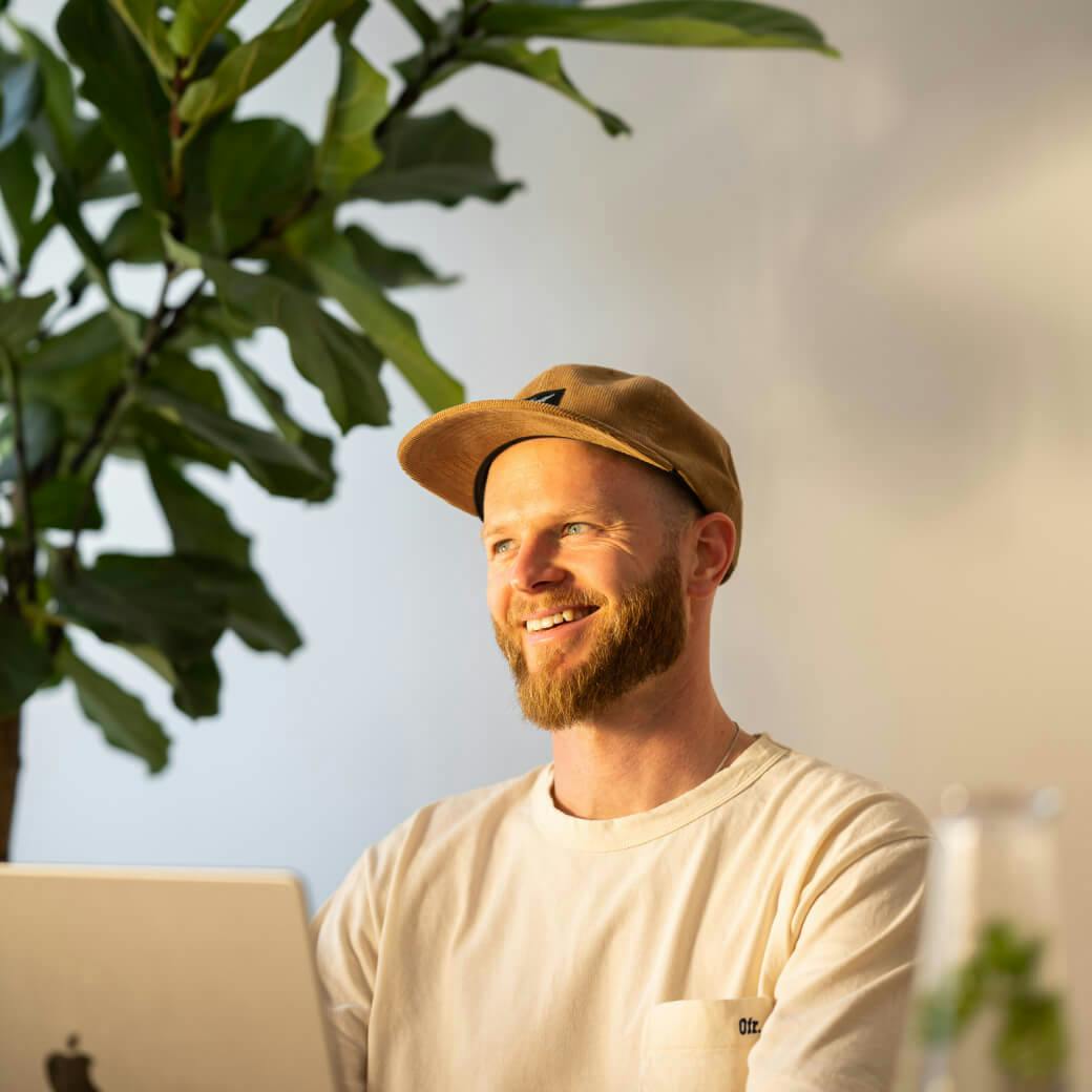 A smiling man in a cap sits next to a laptop, with a plant in the background.