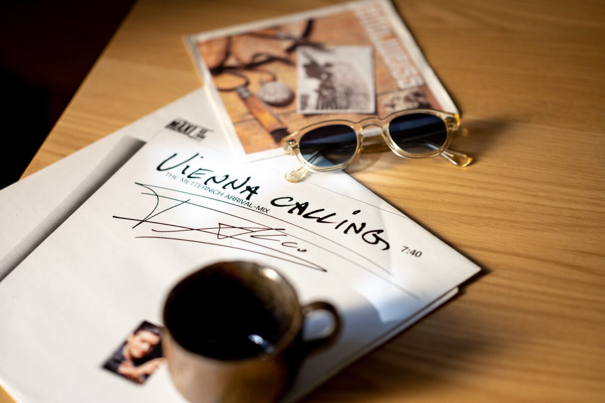 A table with a cup of coffee, a pair of round sunglasses and the Vienna Calling LP by Falco.