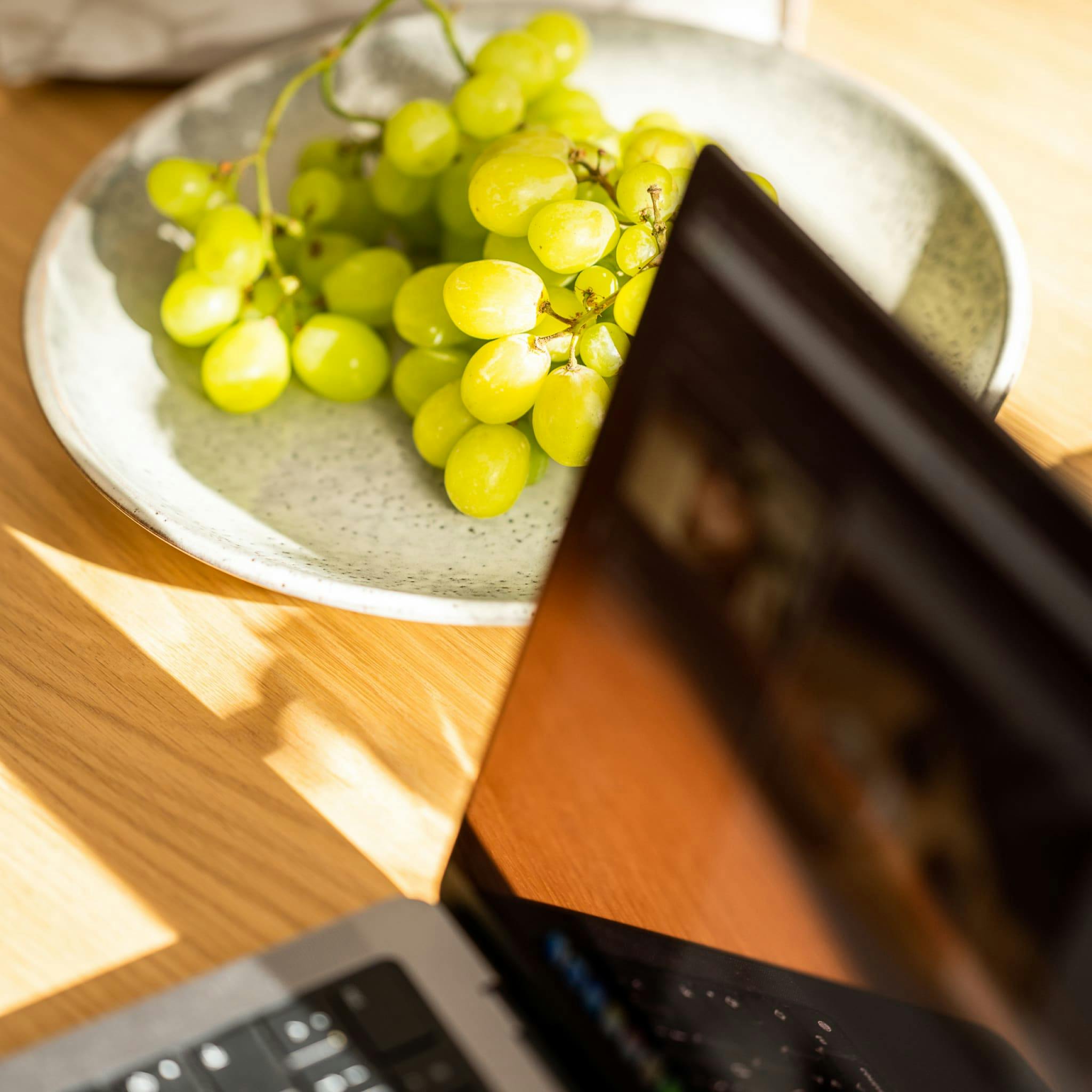 A plate of grapes next to an open laptop on a sunlit wooden table.